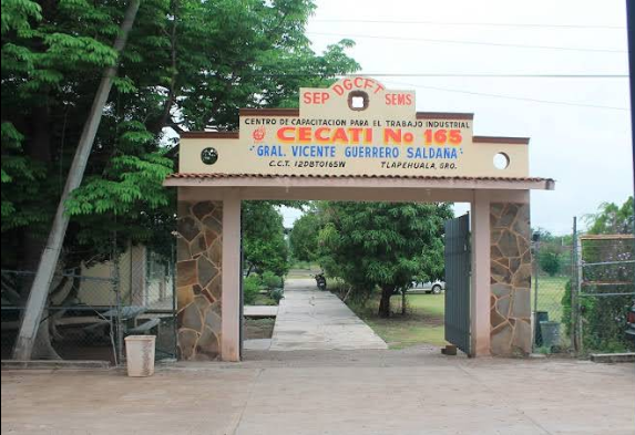 Fotografía en la que se muestra la entrada del plantel, conformada por un arco. En la parte superior del arco dice CECATI NÚMERO 165. Debajo del arco hay una reja de color gris que está abierta, lo que permite el acceso a las instalaciones y se ven las áreas verdes